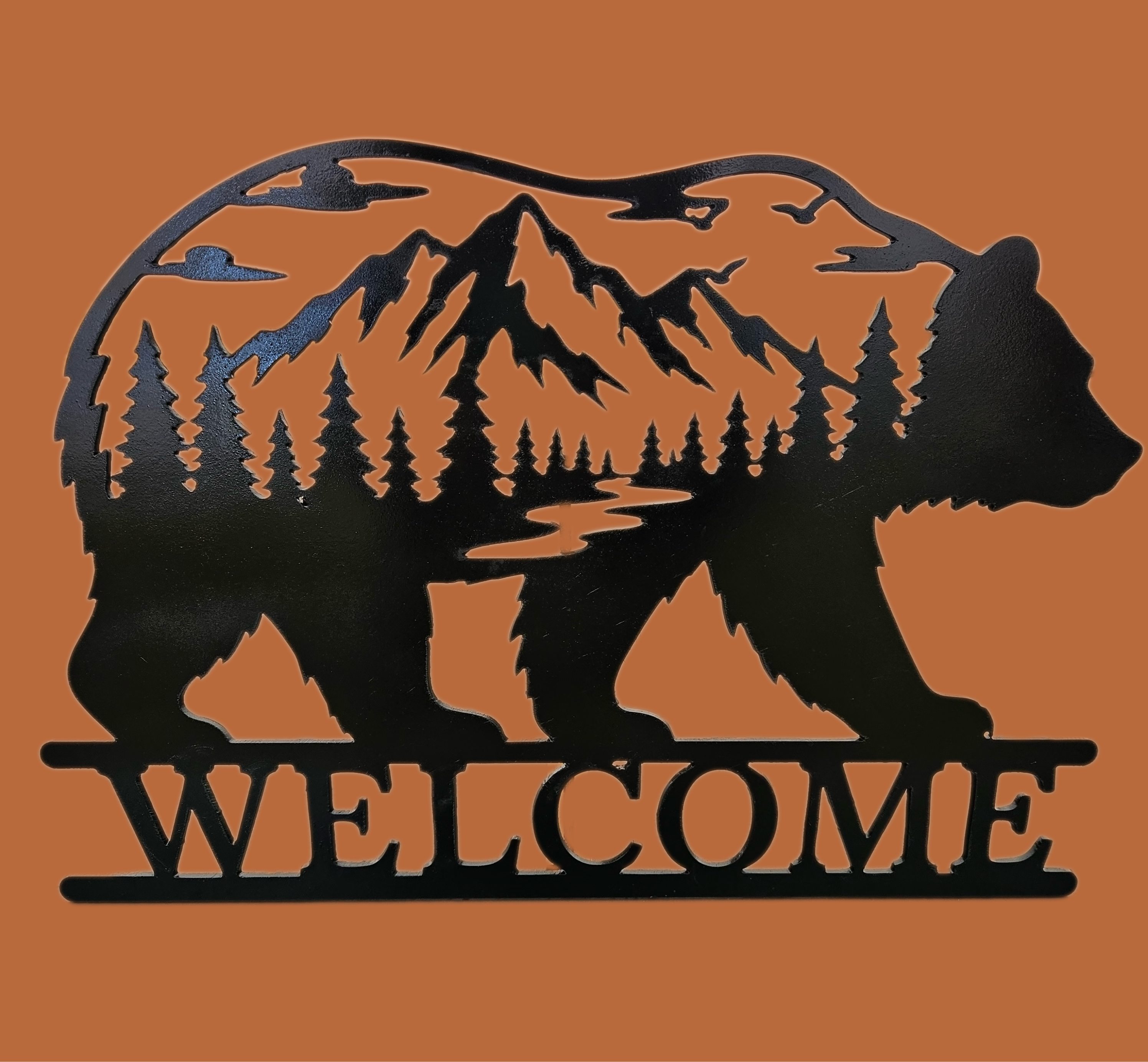 Black metal wall art featuring a bear silhouette filled with a mountain, forest, and river scene, with the word WELCOME across the bottom, shown on a solid orange background.