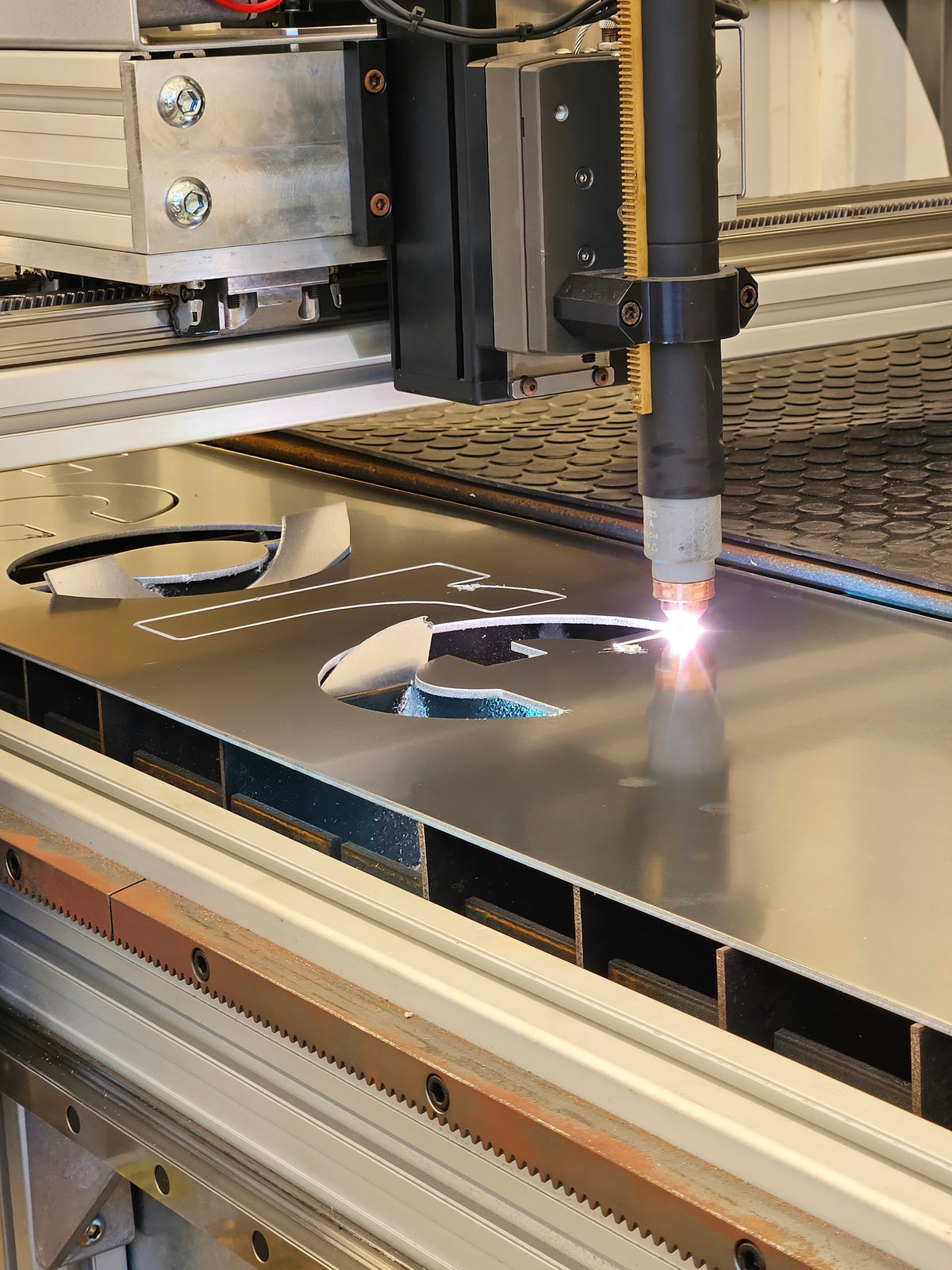 A laser cutting head actively cutting a large sheet of metal, with a bright arc of light visible at the intersection of the nozzle and the metal surface.