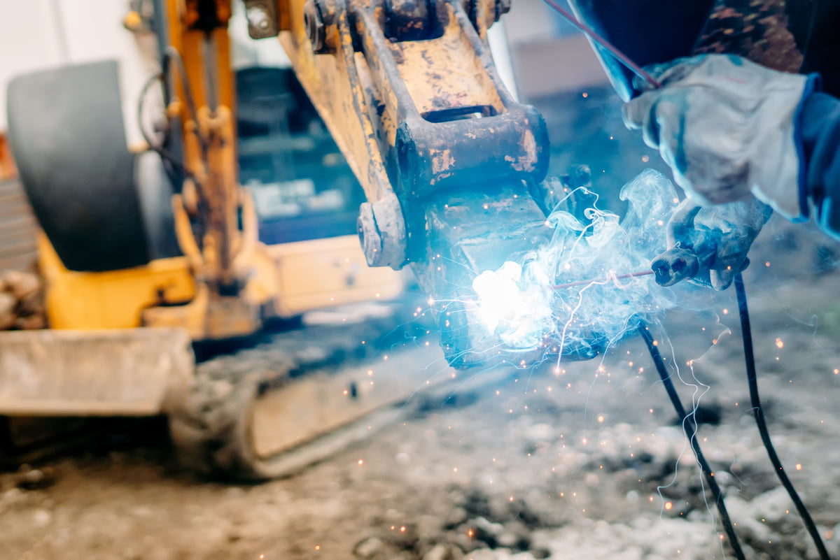 A close-up action shot of a welder in blue protective clothing and gloves holding a welding torch, creating a bright blue-white arc of sparks near a yellow excavator arm in an outdoor work environment.