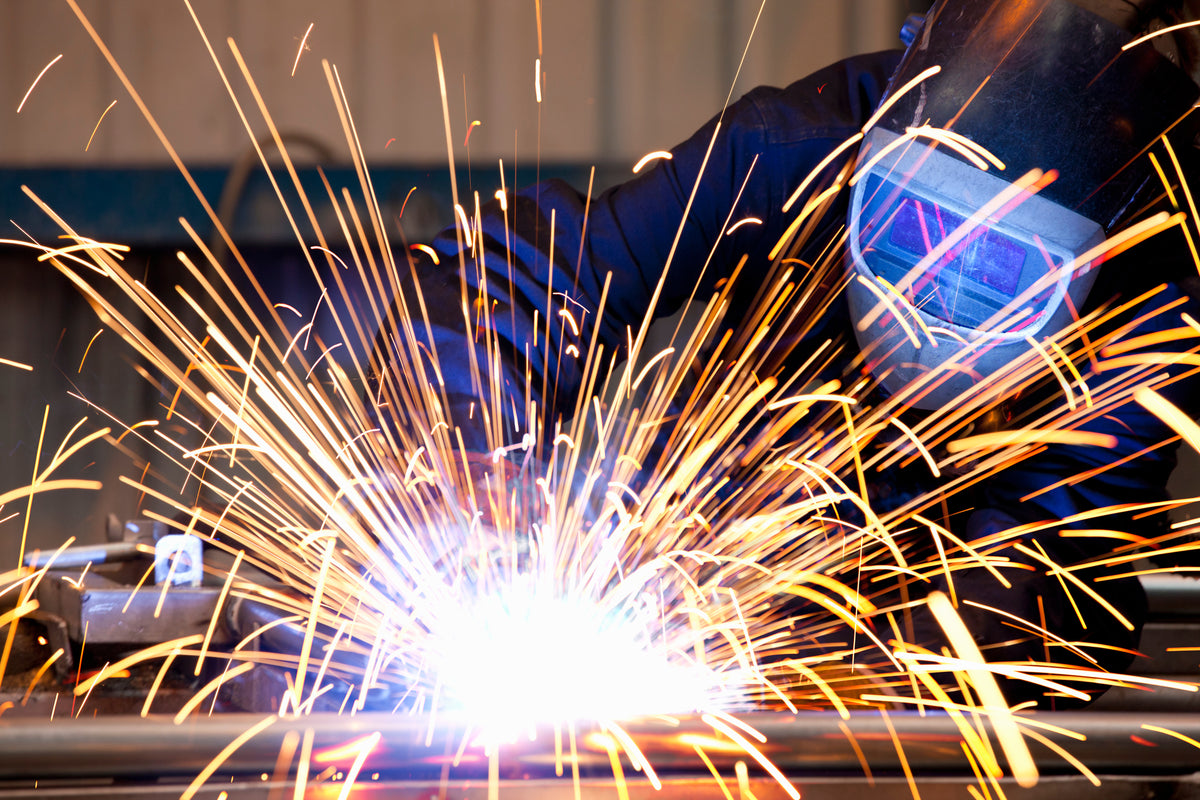 A welder in protective gear wearing a dark helmet generates a bright welding arc with numerous golden-orange sparks flying in all directions against an industrial background.