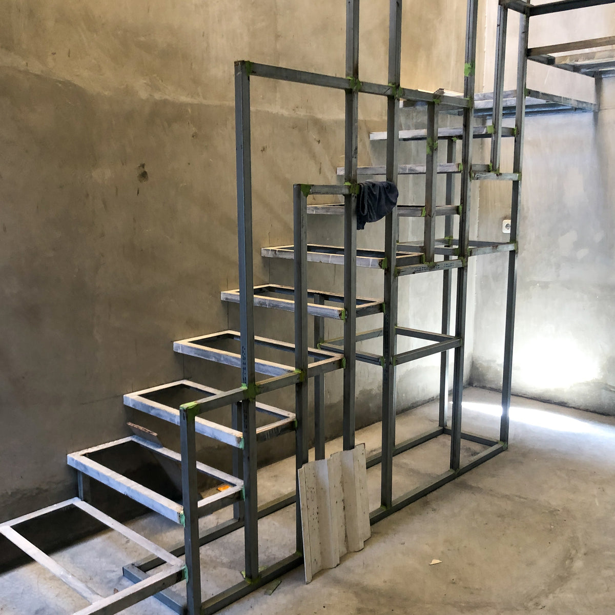 Custom cut metal staircase frame under construction, showing steel steps and handrails in a raw concrete room.